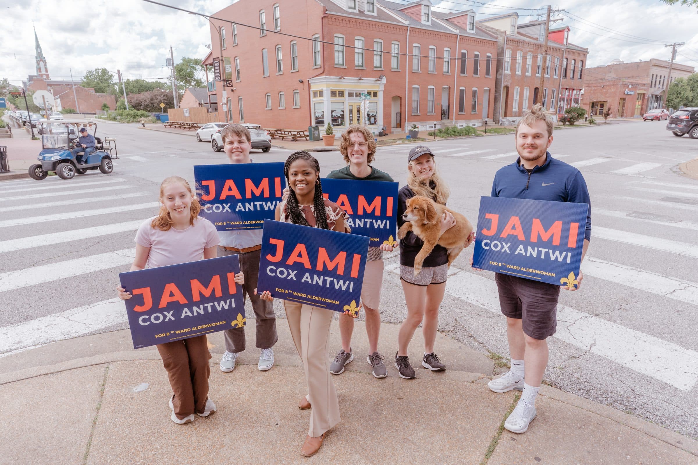 Jami Cox Antwi and volunteers with campaign signs on a crosswalk, brick buildings behind them